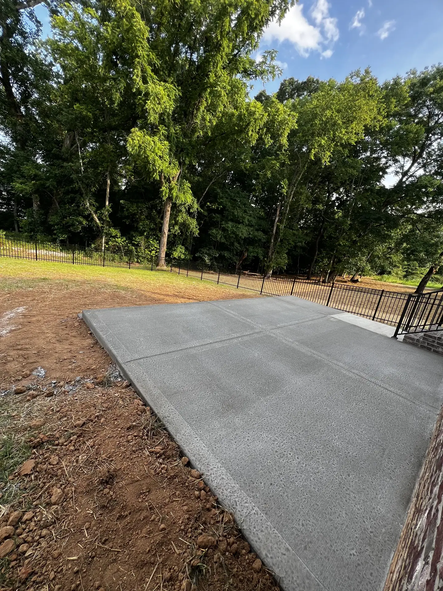 Rectangular concrete patio pad beside black aluminum fencing and trees