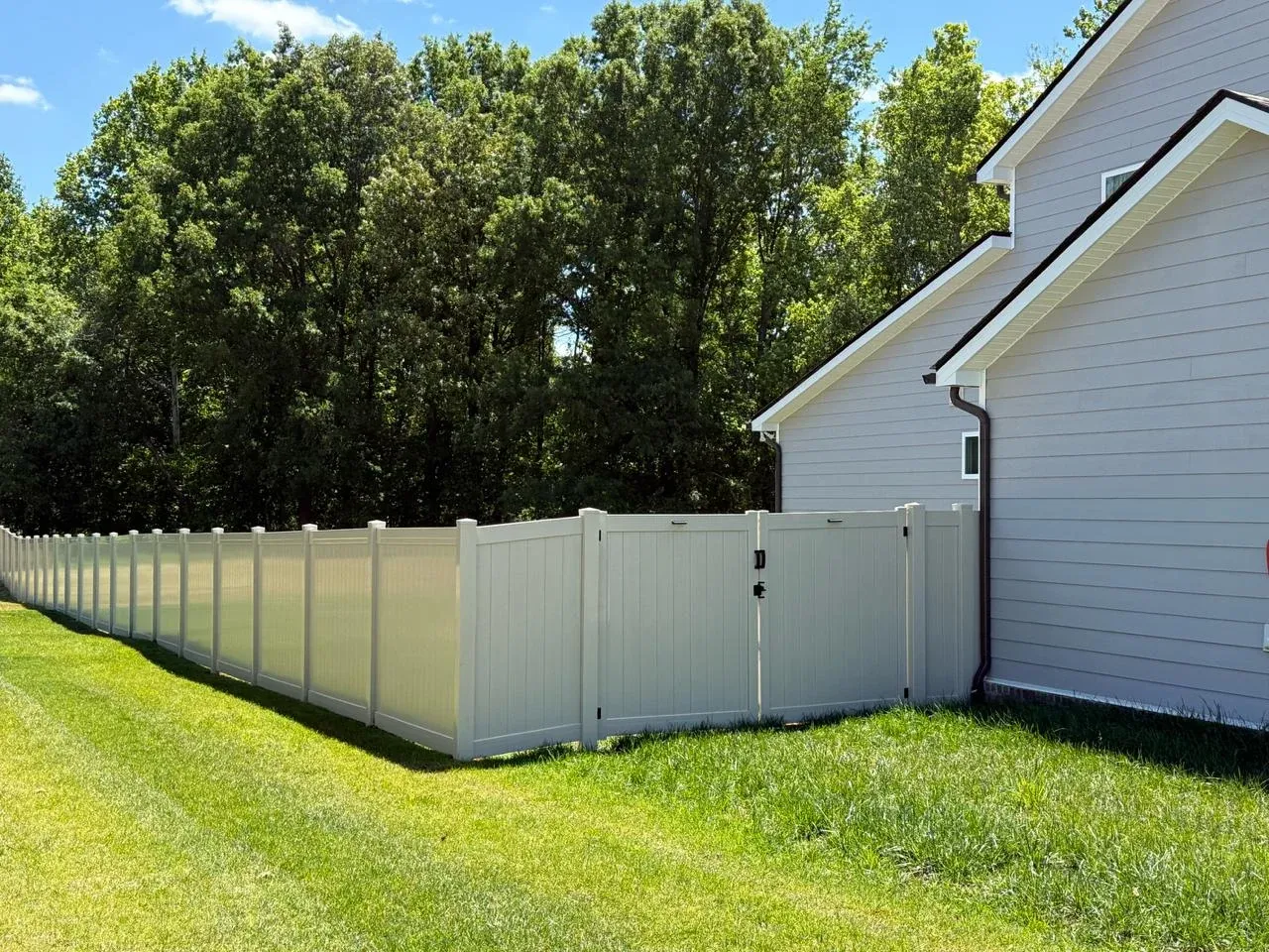 White vinyl privacy fence and gate beside a house and wooded backyard